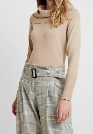 Femme portant un pull beige avec un large col côtelé et un pantalon beige et bleu à carreaux taille haute avec une boucle de ceinture rectangulaire noire.
