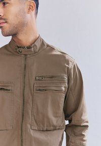 Rjava lahka jakna z visokim ovratnikom, sprednjo zadrgo in dvema žepoma na prsih z zadrgo. Ima gladko teksturo in minimalističen dizajn.