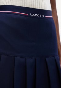 Gros plan d'une jupe plissée bleu marine avec une ceinture élastique arborant le logo Lacoste et des rayures rouges et blanches.