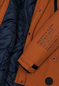 Rękaw kurtki w kolorze rdzy z czarnym napisem „Identity Code Invincible” oraz widoczna granatowa, pikowana podszewka kurtki.