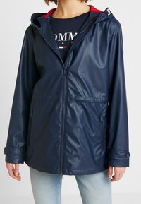 Laivansininen vedenpitävä takki, jossa on vetoketjukiinnitys, säädettävä vyötärö ja punainen huppu. Takissa on kaksi sivutaskua ja nappikiinnitteiset hihansuut.