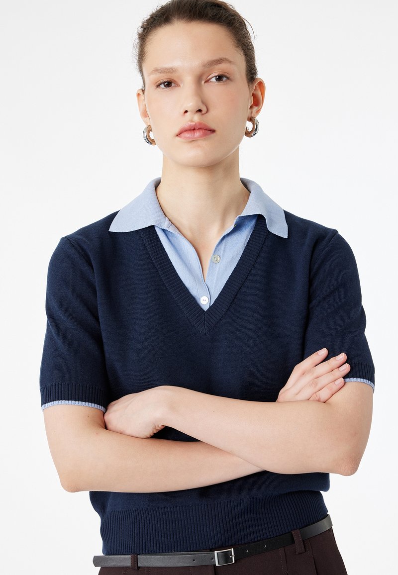 Ung kvinde med armene krydsede iført en marineblå V-hals sweater over en lyseblå skjorte med krave og sølvfarvede øreringe, neutral baggrund.