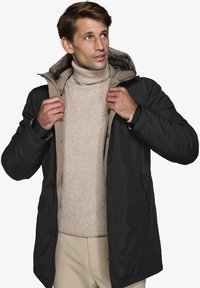 Giacca nera isolante con fodera beige, dotata di zip e cappuccio regolabile. Indossata sopra un maglione a collo alto beige chiaro.