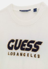 Valge koobatud sviiter, millel on soonikuga ümmargune kaelus. Esiküljel on tume sinine "GUESS" ja kuldne "LOS ANGELES" tikand.