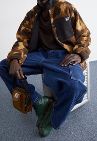 Hombre sentado con chaqueta de forro polar marrón y negra, vaqueros azules holgados, botas verdes y bolso cruzado marrón de Carhartt.