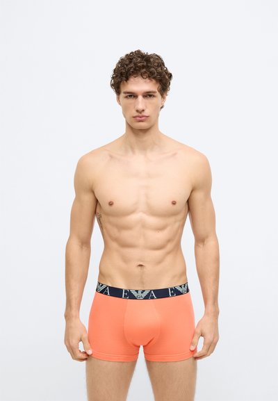 Boxers largos laranja feitos de tecido macio, com uma cintura de cor azul escura e um logotipo. O design é justo, com um bolso contornado.