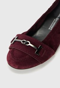 Scarpa piatta in suede borgogna con punta arrotondata, dotata di una fascia in metallo nero con dettaglio del logo e cuciture visibili lungo il bordo.