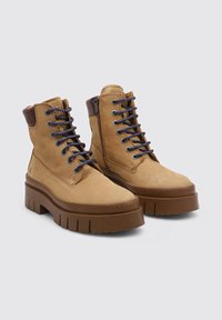 Botas de tobillo de cuero color camel con suelas gruesas de color marrón, cuellos acolchados marrones oscuros, cordones oscuros y cremalleras laterales sobre un fondo sencillo.