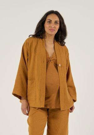 Zwangere vrouw met donker haar draagt een los, getextureerd mosterdkleurig outfit met een jasje met knopen en bijpassende broek, staand tegen een effen achtergrond.