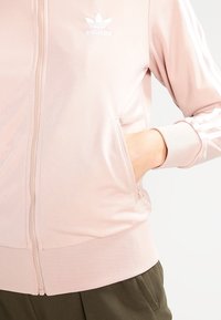 Růžová bunda na zip se žebrovanými manžetami a lemem, vybavená bílým logem Adidas a boční kapsou, vyrobená z hladkého, lehkého materiálu.