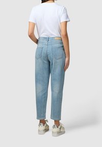 Žena v světlemodrých vysokých džínech, bílé košili s krátkým rukávem a bílých teniskách se stříbrnými detaily, stojící na jednobarevném pozadí.