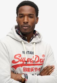 Grijze hoodie met rode "SuperDry"-logo, witte trekkoorden, boven een geruit overhemd. Opvallend horloge om de pols, neutrale achtergrond.