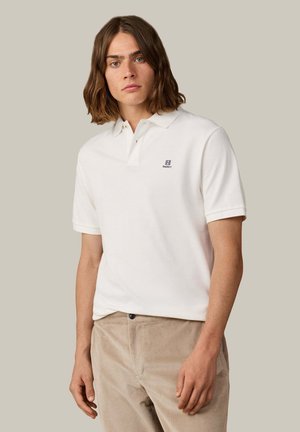 Ung mand med skulderlangt hår iført en hvid polo-shirt og beige kordfløjlsbukser mod en ensfarvet baggrund.