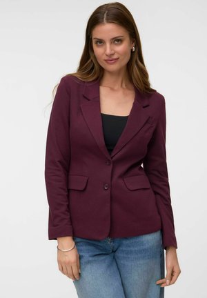 Femme aux longs cheveux bruns portant un blazer violet foncé, un haut noir, un jean bleu et des boucles d'oreilles créoles argentées sur fond uni.