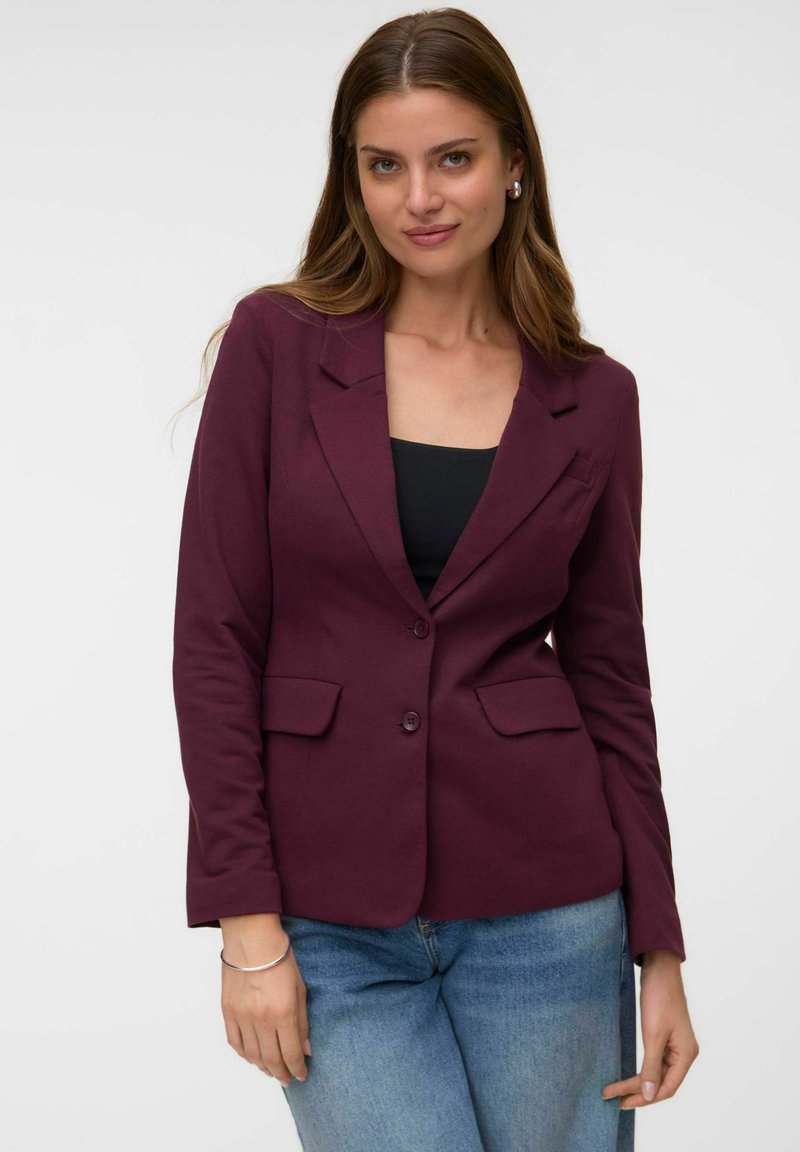 Femme aux longs cheveux bruns portant un blazer violet foncé, un haut noir, un jean bleu et des boucles d'oreilles créoles argentées sur fond uni.