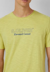 Jasnożółta bawełniana koszulka z okrągłym dekoltem. Posiada nadrukowane logo "S.OLIVER" oraz napis "Elevated Casual" w ciemnoniebieskim kolorze. Gładka tekstura.
