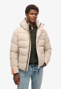 Beige pufferjakke med hætte, med lynlås-lukning, sorte accenter og et mærke på ærmet. Bæres over en mørkegrøn sweater og jeans.