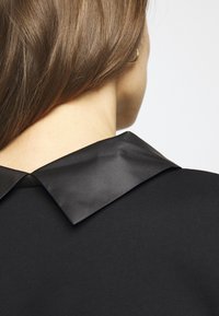 Černé šaty s lesklým, širokým límečkem. Textura kontrastuje s matnou tkaninou. Vlasy padají přes rameno a odhalují zlatý náušník.