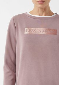 Jasnoróżowy sweter z teksturowanym prostokątnym logo w kolorze różowego złota z napisem "Mniej znaczy więcej". Gładka tkanina i okrągły dekolt.