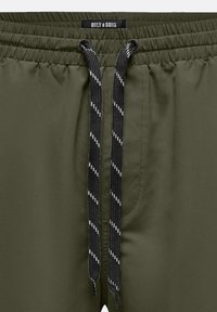 Pantalones cortos verde oliva con cintura elástica, que presentan cordones negros con rayas blancas. Textura suave y detalles de diseño mínimos.