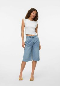 Biały krótki top z koronkowymi akcentowanymi rękawami, zestawiony z jasnoniebieskimi dżinsowymi culottes. Modelka ma na sobie żółte sandały na obcasie.