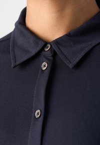 Gros plan d'une chemise bleu marine foncé à col avec trois boutons gris visibles fermés au niveau du cou et du haut de la poitrine d'une personne.