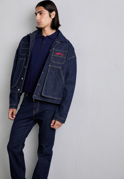 Denim jacket featuring a button closure, two front pockets, and red logo embroidery; paired with dark denim pants and a navy polo shirt.