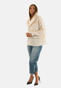 Frau in einem cremefarbenen, oversized Strickpullover mit umgeschlagenen Bündchen, hellblauen, hochgekrempelten Jeans und schwarzen High Heels, die seitlich steht.