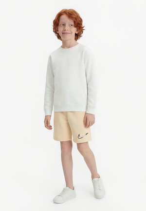 Raudonplaukis berniukas dėvi baltą džemperį, smėlio spalvos „Nike“ šortus ir baltus sportbačius, stovi ir žiūri į šoną, šiek tiek šypsodamasis.