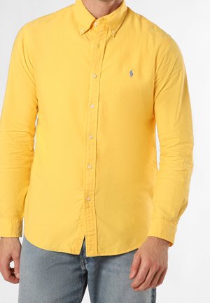 Homme portant une chemise boutonnée jaune vif à manches longues avec col et un jean bleu clair, debout devant un fond blanc uni.