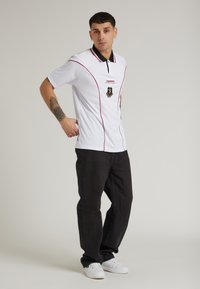 Hvid polo skjorte med sort krave og røde sting, prydet med et logo på brystet. Sammen med sorte jeans og hvide sneakers.