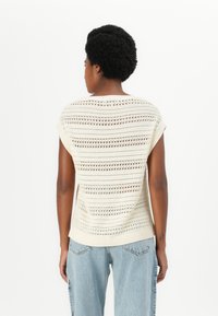 Kremfarget strikket topp med korte ermer som har et åpent mønster med horisontale striper. Myk tekstur og løs passform, kombinert med lyseblå jeans.