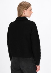 Maglione nero lavorato a maglia con collo alto, tessuto strutturato e design cropped. Abbinato a jeans grigi scuri a vita alta, che mostrano un look minimalista.