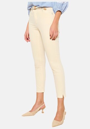 Jean skinny couleur crème avec une texture lisse, fentes aux chevilles et taille mi-haute. Associé à des talons aiguilles beiges.
