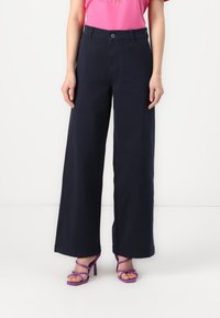 Calças largas de cor navy, feitas de algodão, com fecho de botão e bolsos frontais. Combinadas com saltos altos roxos com tiras.