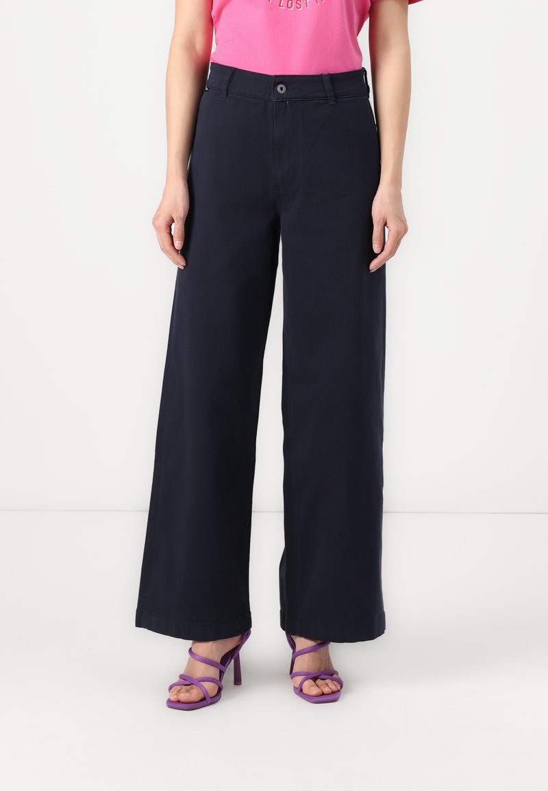 Calças largas de cor navy, feitas de algodão, com fecho de botão e bolsos frontais. Combinadas com saltos altos roxos com tiras.