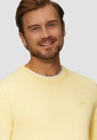 Gelb gestrickter Pullover mit rundem Halsausschnitt und einem dezenten Logo auf der Brust, mit gerippten Bündchen und Saum, weiche Textur.