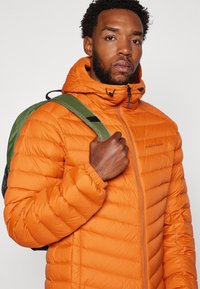 Puffermantel i lys orange med quiltet design, justerbar hætte og lynlås lukning. Bæreren holder en grøn rygsækkelukning på venstre skulder.