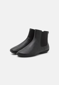 Botas de cuero negro hasta el tobillo con una textura suave, paneles laterales elásticos y punta redondeada. Suela plana con detalles mínimos.