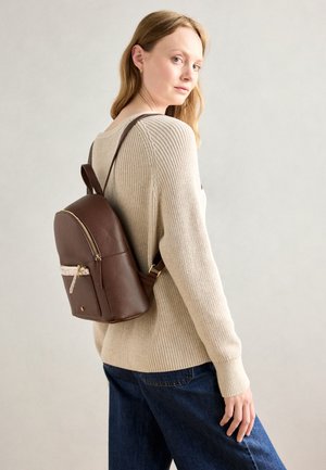 Sac à dos en cuir marron avec une forme arrondie, fermeture éclair dorée et une poche avant. Le modèle porte un pull crème et un jean en denim bleu.