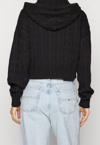 Back view of person wearing black cable-knit hooded sweater and light blue high-waisted jeans with back pockets and small logo.