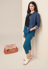 Camicia di jeans, top nero, pantaloni cropped teal e sneakers bianche con dettagli beige. Una grande borsa bicolore arancione e rosa è sul pavimento.