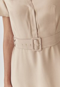 Beiges Kleid mit einem taillierten Gürtel und einer quadratischen Schnalle sowie subtilen Falten, aus glattem Stoff mit weicher Textur.
