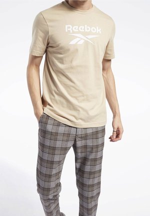 Homme portant un t-shirt beige Reebok et un pantalon à carreaux gris, les mains dans les poches, sur fond blanc.