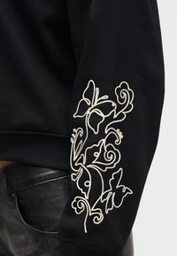 Manche de sweat-shirt noire avec motifs floraux et papillons brodés en blanc au-dessus d'un pantalon en cuir noir.