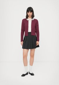 Cardigan côtelé bordeaux avec des boutons, chemise à col blanc, jupe plissée grise, chaussettes blanches hautes aux genoux et ballerines noires.