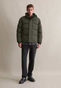 Jachetă puffer de culoare verde închis, cu secțiuni cusute și guler înalt. Purtați-o peste o cămașă în carouri, asociată cu blugi închisi și pantofi negri.