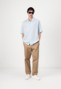 Camicia azzurra a maniche corte con bottoni e vestibilità relaxed, abbinata a pantaloni beige e sneakers bianche, indossata con occhiali da sole scuri.