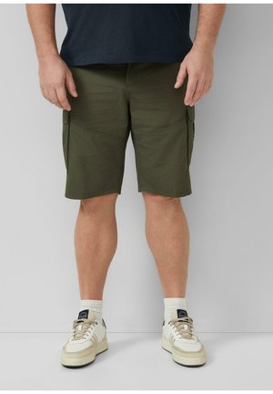 Man draagt donkergroene cargoshorts, witte sokken en witte sneakers en staat tegen een effen lichtgrijze achtergrond.