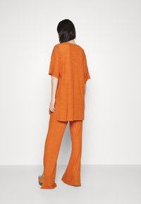 Henkilö seisoo selkä kameraan päin, päällään löysä ribattua oranssia toppia ja siihen sopivat leveälahkeiset housut, yksinkertaista valkoista taustaa vasten.